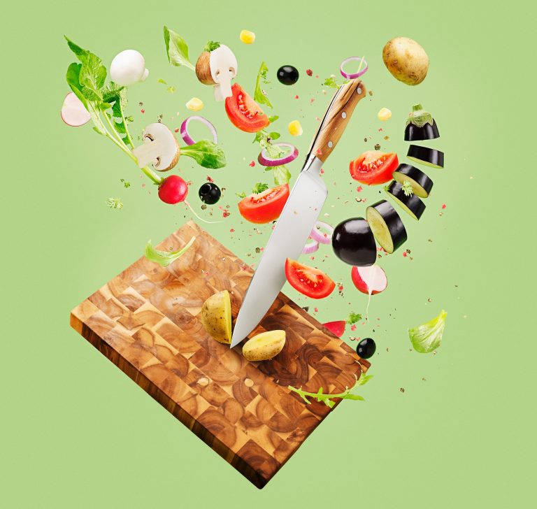 Das Bild zeigt ein Schneidebrett auf dem ein schwendes Messer steht, welches von vielen verschiedenen geschnittenen Obst, Gemüse und Kräutern umgeben ist - eine Art Strudel entsteht, der sich auf die Schneide des Messers fokussiert - Das Motiv wurde vom Studio Hackenberg in Hamburg für den Kunden ChefsCulinar produziert, um als Titelmotiv des Obst und Gemüsekatalogs verwendet zu werden