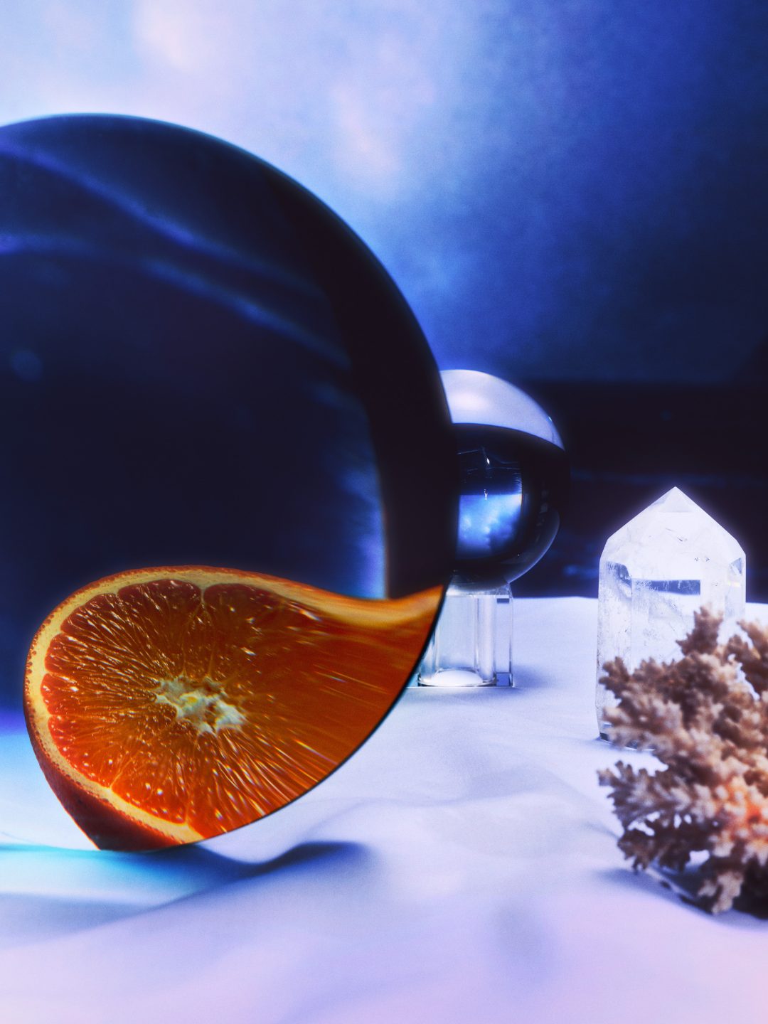 Foto eines Tisches mit diversen Objekten darauf liegend, etwa einer Orange die durch eine Lupe verzerrt wird, in einem stimmungsvollen traumähnlichen Setting, getaucht in Lila-blaues Licht - Foto der Strecke Lunatismus des Studio hackenberg