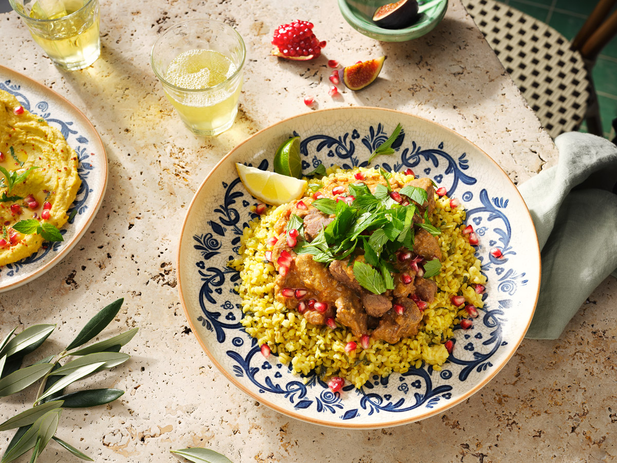 Foodfotografie von Studio Hackenberg in Hamburg: Entdecken Sie unser Bild eines Bulgur-Lammragouts in mediterranen setting angerichtet mit gelbem Bulgur, Granatapfelkernen und frischen Kräutern, umgeben von Hummus und Feigen.