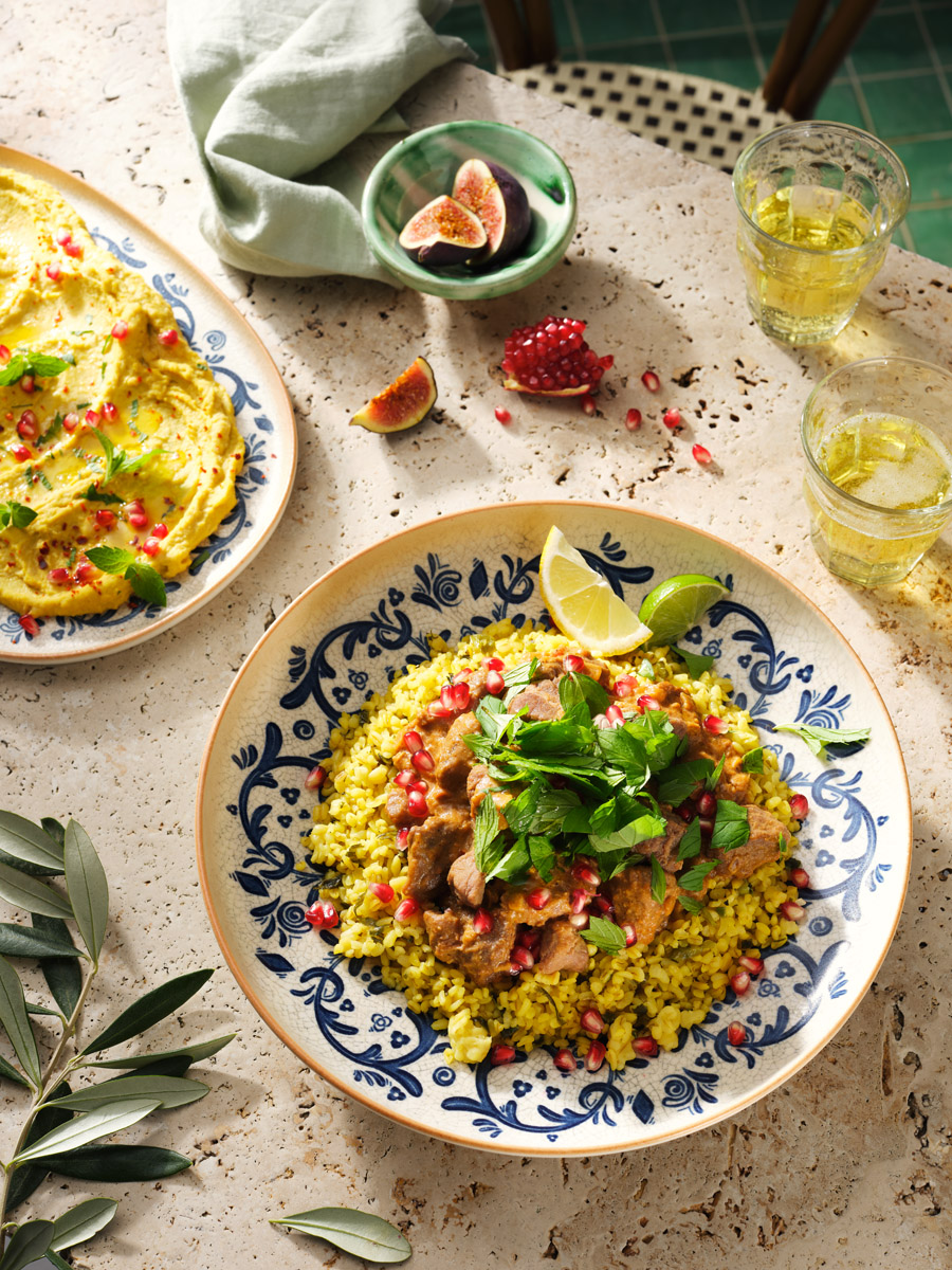Einblick in die Foodfotografie von Studio Hackenberg in Hamburg: Entdecken Sie unser Bild eines Bulgur-Lammragouts, angerichtet mit gelbem Bulgur, Granatapfelkernen und frischen Kräutern, umgeben von Hummus und Feigen.